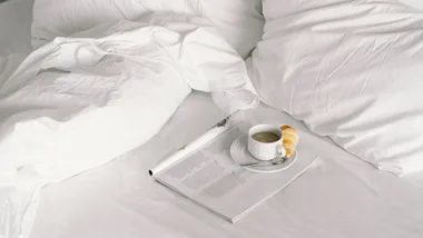 Unmade bed with white cotton sheets and a coffee on a magazine