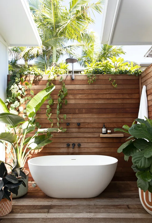 Outdoor bathroom with white tub, surrounded by lush plants and wooden walls, under a blue sky.