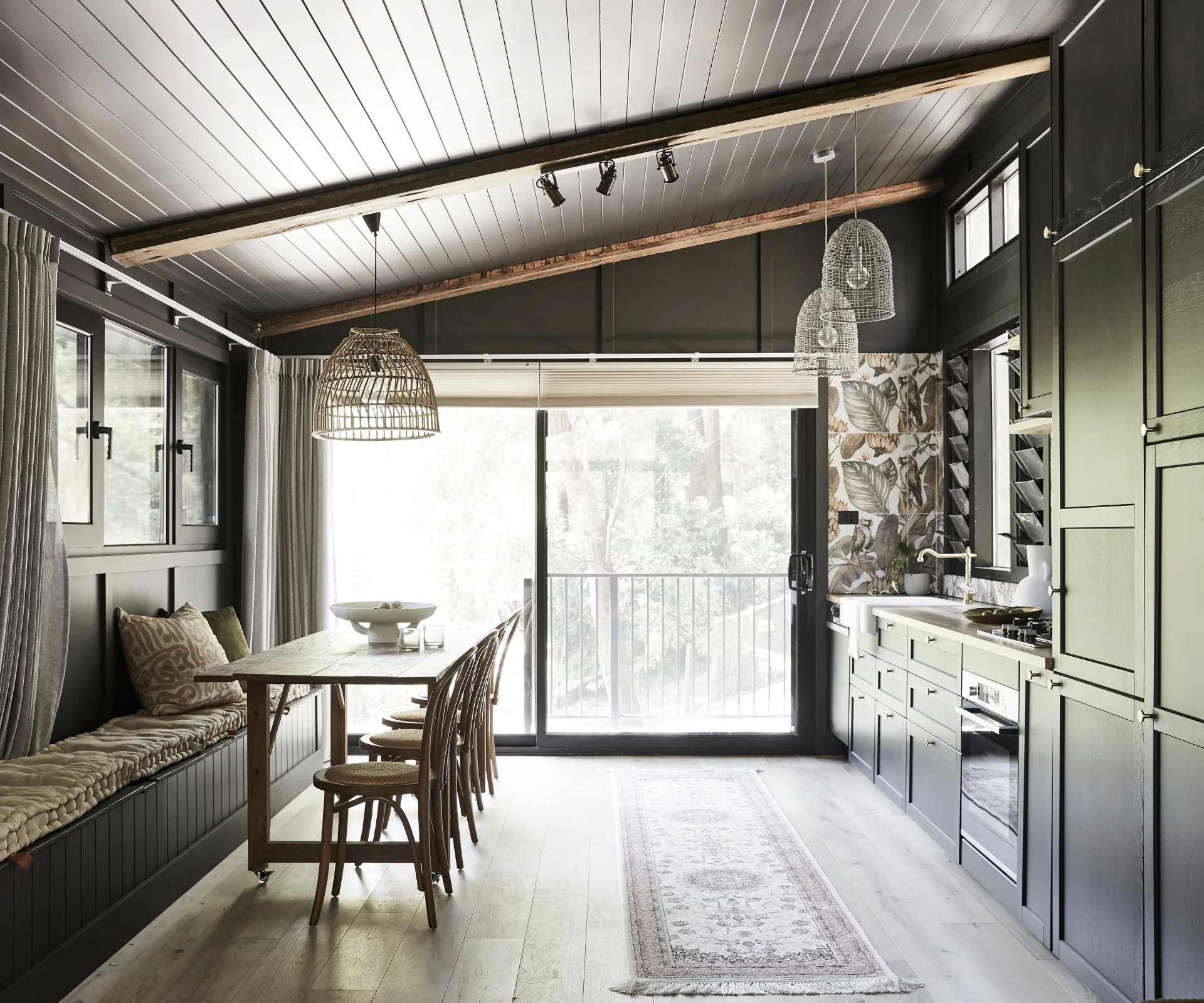 A small black granny flat with a kitchen area one side and a dining area on the other with raw timber table and bench seating.