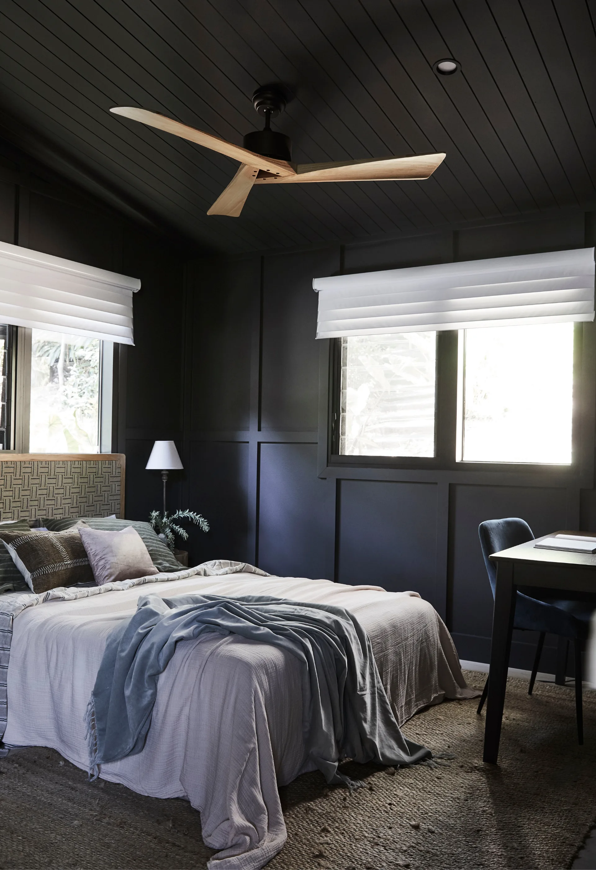 A bedroom with black walls and ceiling, with light-timber ceiling fan and bed with white and blue linens