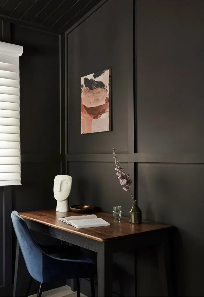 An all-black room with a polished timber desk topped with a single flower and a white face sculpture. A blue velvet chair is tucked into the desk, and a small pink abstract artwork hangs on the walls.