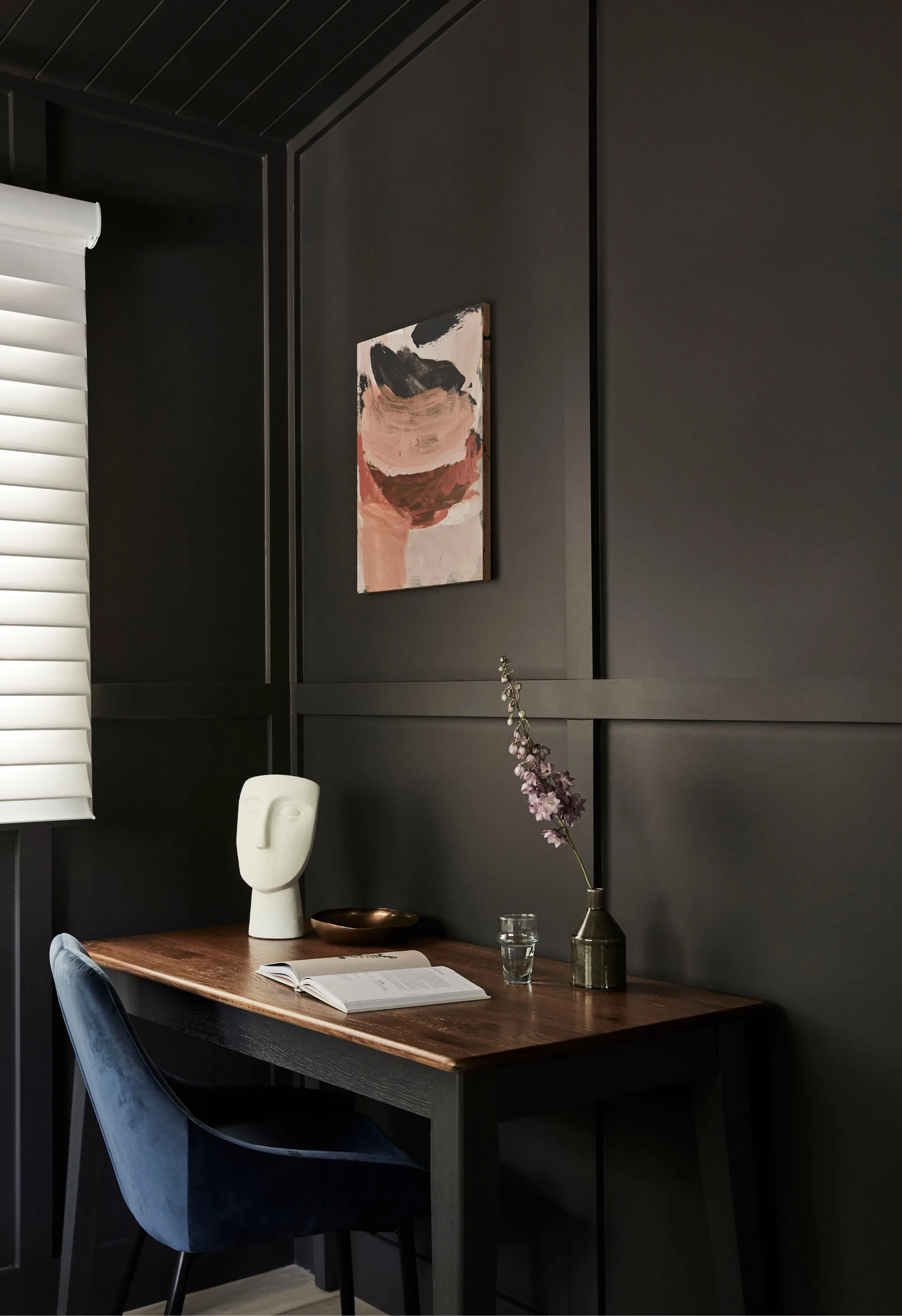 An all-black room with a polished timber desk topped with a single flower and a white face sculpture. A blue velvet chair is tucked into the desk, and a small pink abstract artwork hangs on the walls.