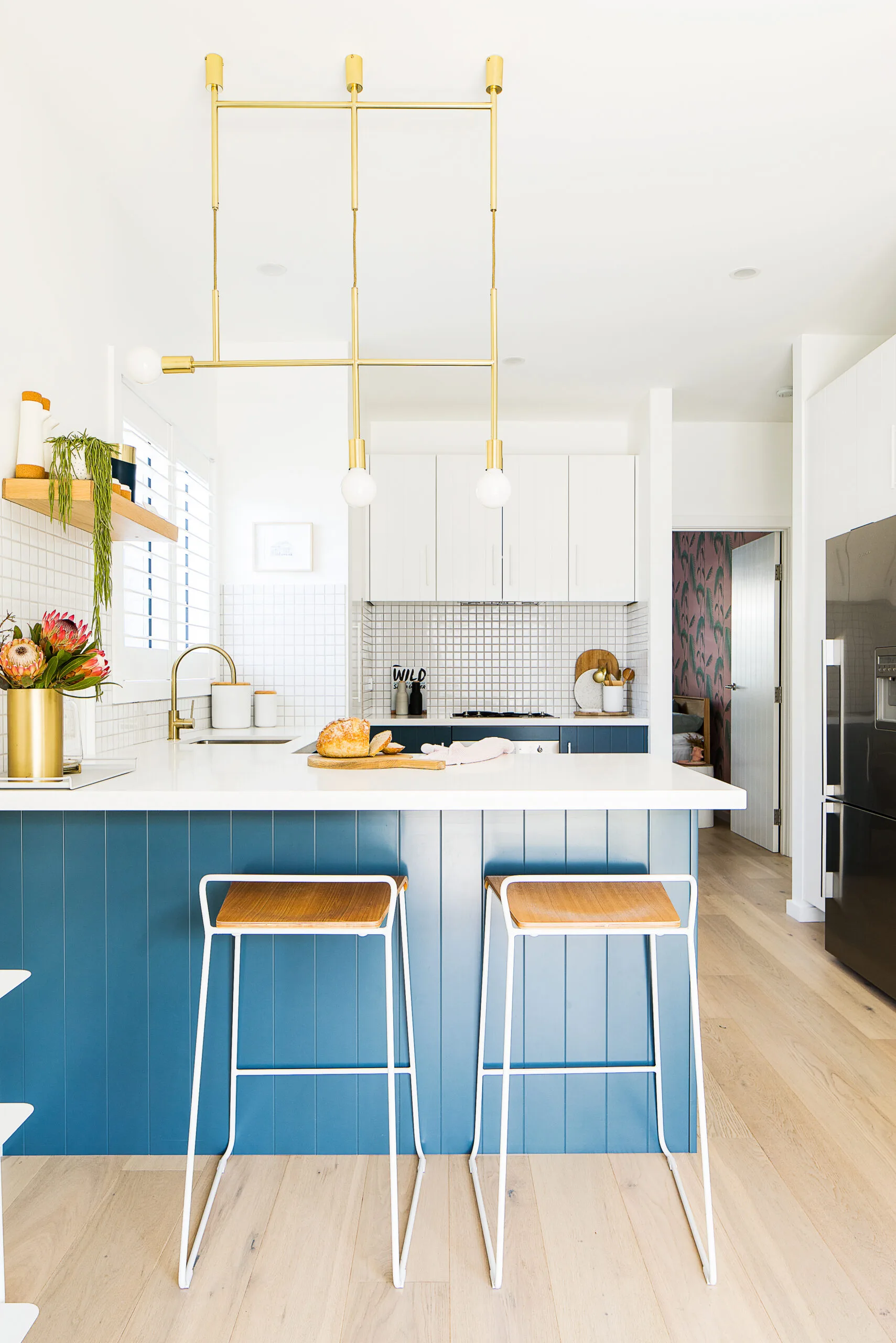 Modern kitchen with blue island, two stools, gold pendant lights, and countertop with bread and flowers.