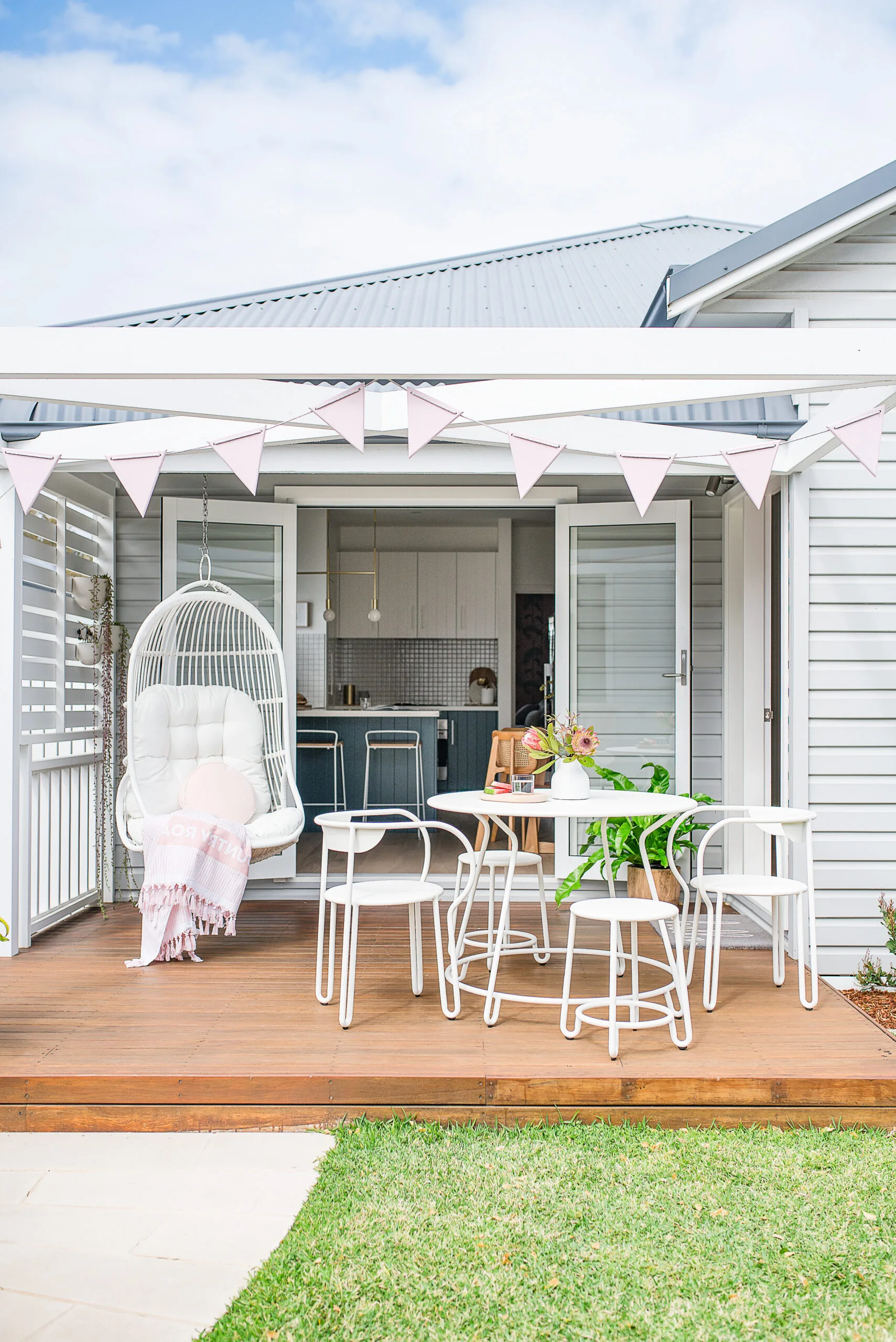 The exterior of a light-grey, weatherboard granny flat with double doors and a small timber deck with white outdoor furniture