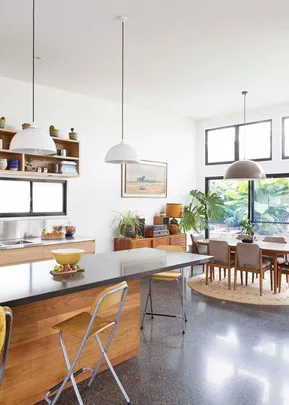 Modern kitchen and dining area with large windows, plants, hanging lights, and wooden furniture, featuring a sleek black countertop.