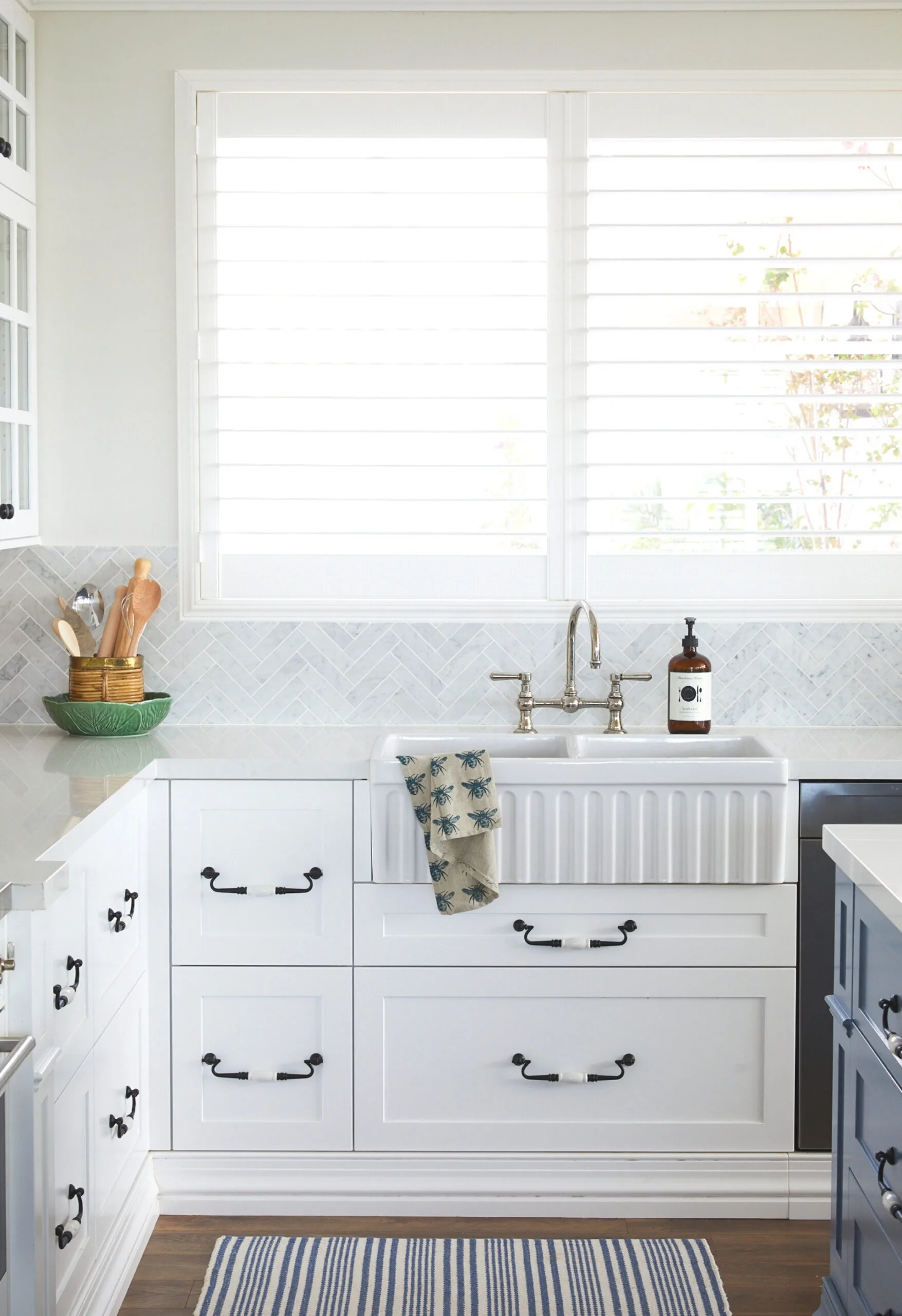 Bright kitchen with white cabinets, farmhouse sink, striped rug, and window with shutters.