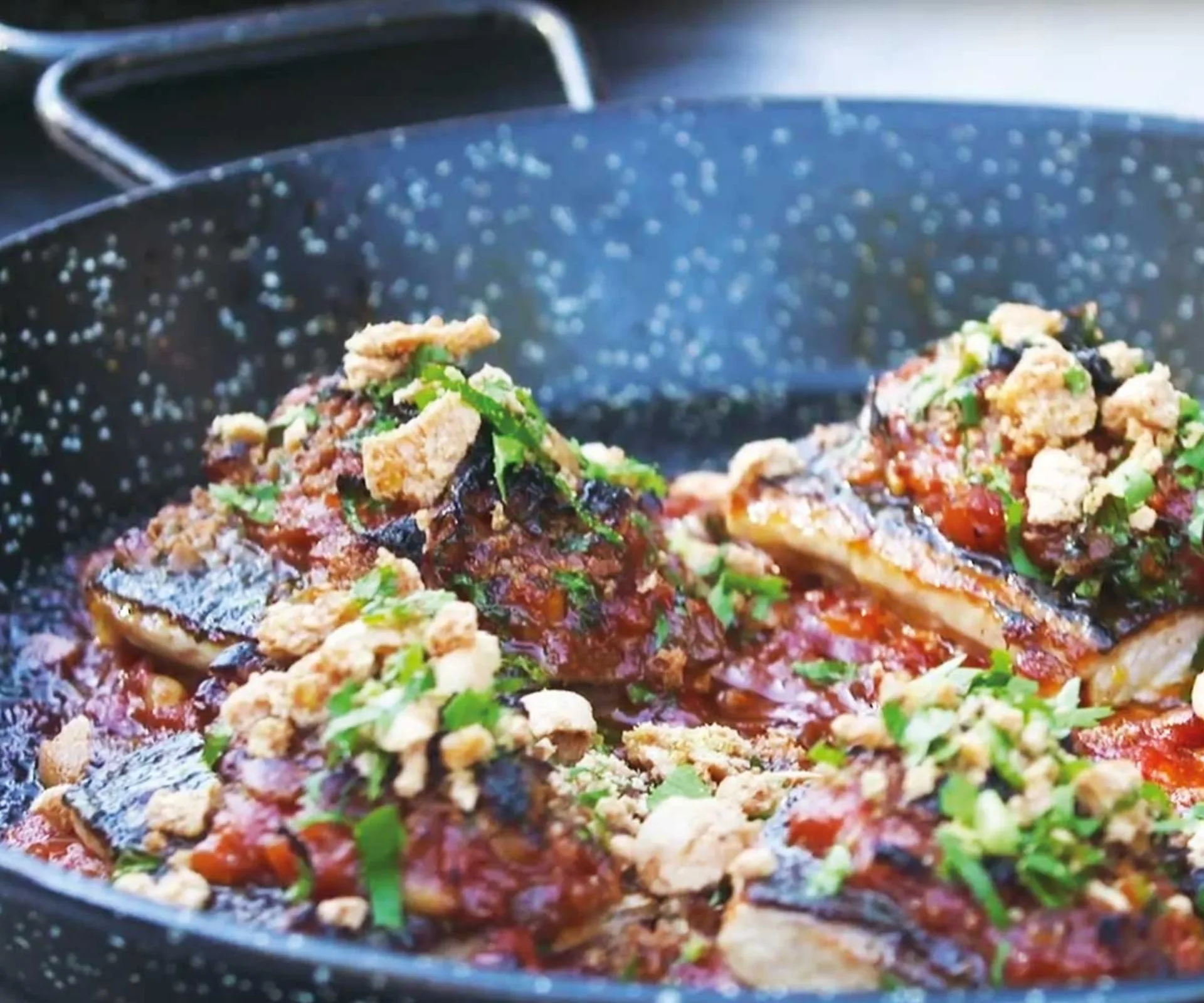 Sizzling fish topped with herbs, sauce, and crunchy crumbs in a speckled pan.