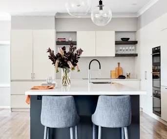 Modern kitchen with a white island, two gray chairs, a vase of flowers, and pendant lights.