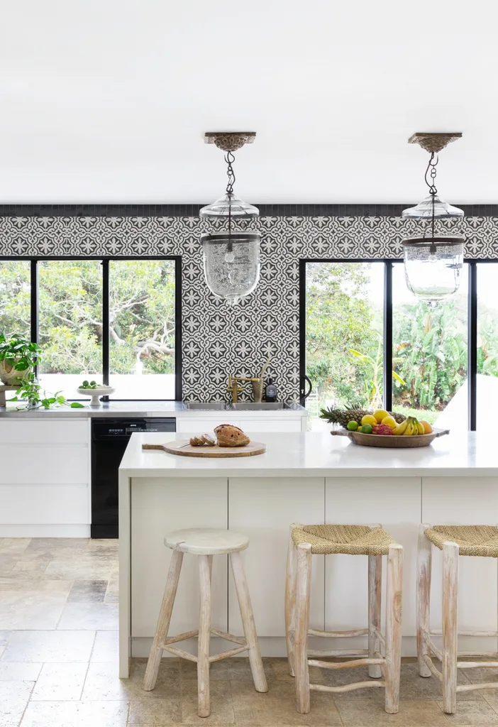 Modern kitchen with patterned tiles, white island, wooden stools, pendant lights, and a view of greenery outside.