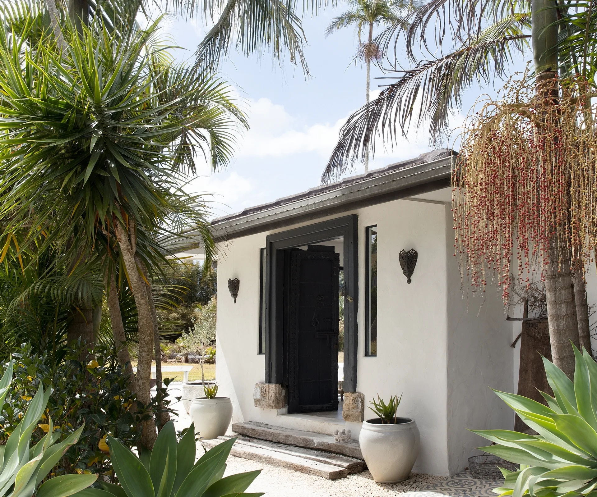 Small white building with a black door, surrounded by tropical plants and palm trees under a clear blue sky.
