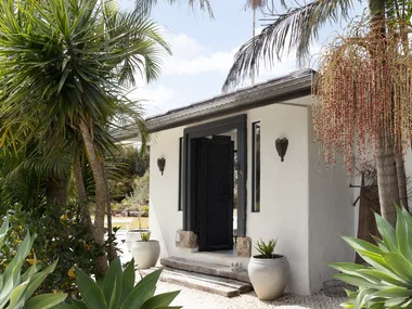 Small white building with a black door, surrounded by tropical plants and palm trees under a clear blue sky.