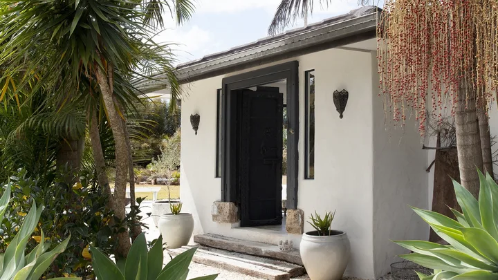 Small white building with a black door, surrounded by tropical plants and palm trees under a clear blue sky.