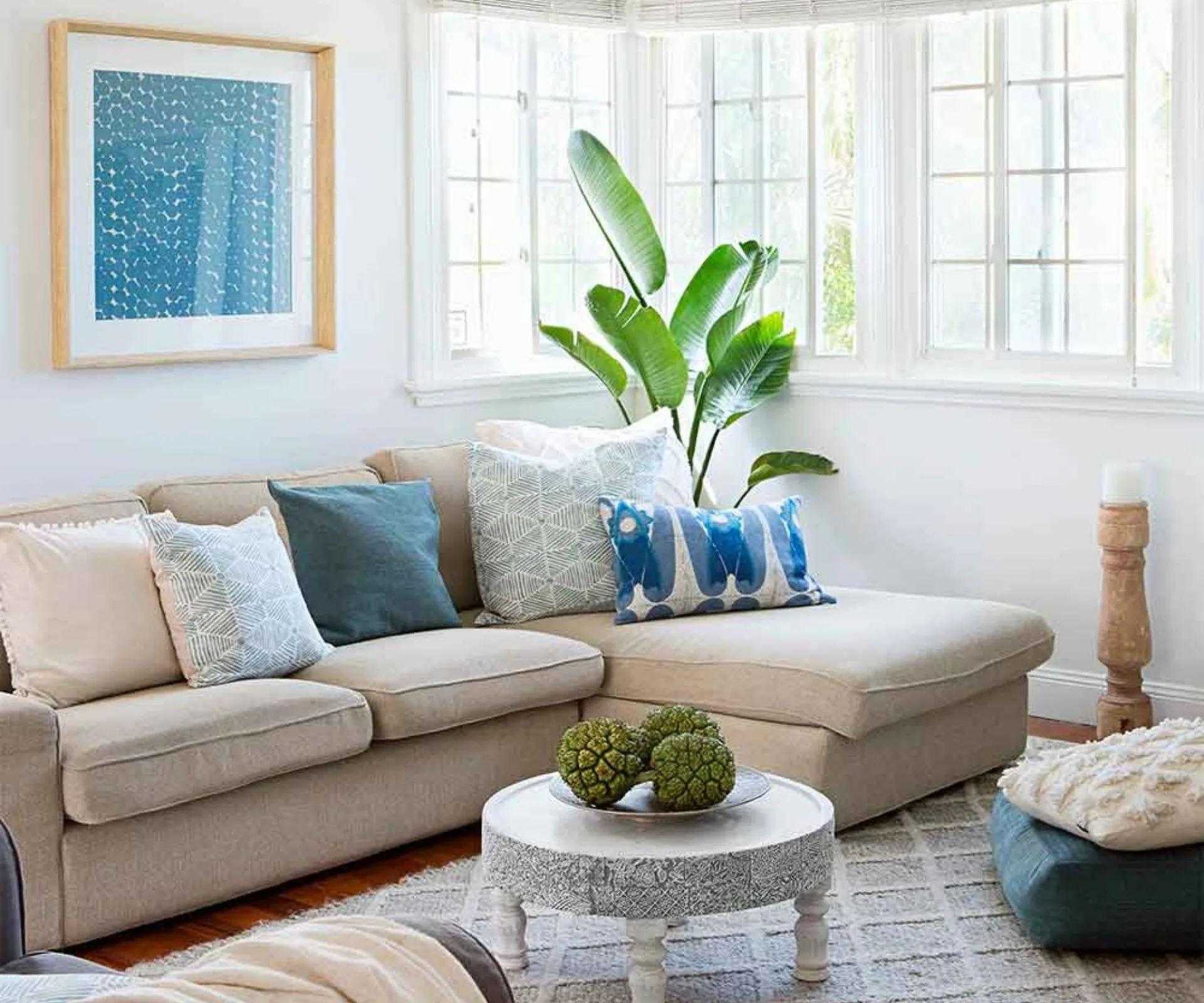 Beige sectional sofa with blue and white pillows, indoor plant, art piece, and round coffee table in bright room.