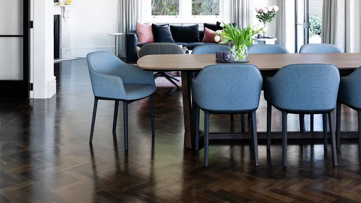Dining room with a large wooden table, blue chairs, pendant lights, and a view of curtains and greenery through windows.