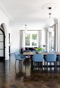 Dining room with a large wooden table, blue chairs, pendant lights, and a view of curtains and greenery through windows.