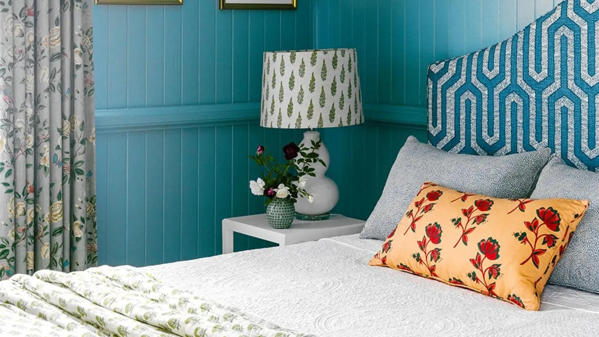 Cozy bedroom with blue walls, floral curtains, patterned lamp, floral pillows on bed, and white side table with flowers.