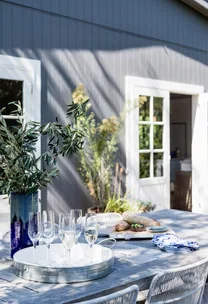 Outdoor table with a vase of greenery, glasses of champagne, bread, and an open patio door in sunlight.