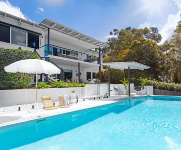 Modern white house with large pool, patio, sun loungers, and umbrellas, surrounded by trees under a clear blue sky.