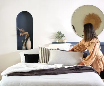 A woman in a beige dress arranges pillows on a neatly made bed with a mirror and decor on the wall behind.