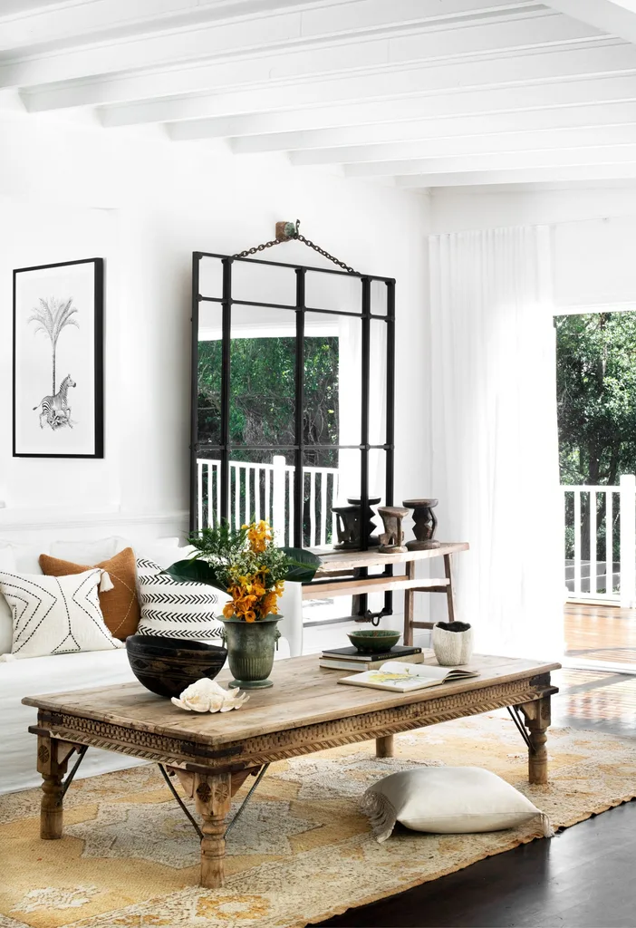 Cozy living room with a rustic wooden table, white sofa, patterned pillows, and large window mirror reflecting greenery.