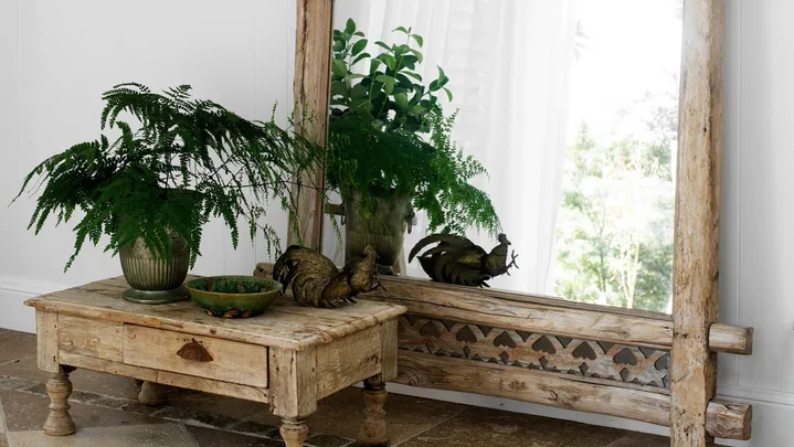 Rustic mirror with green panel beside table holding plants and rooster figurine, on checkered tile floor.