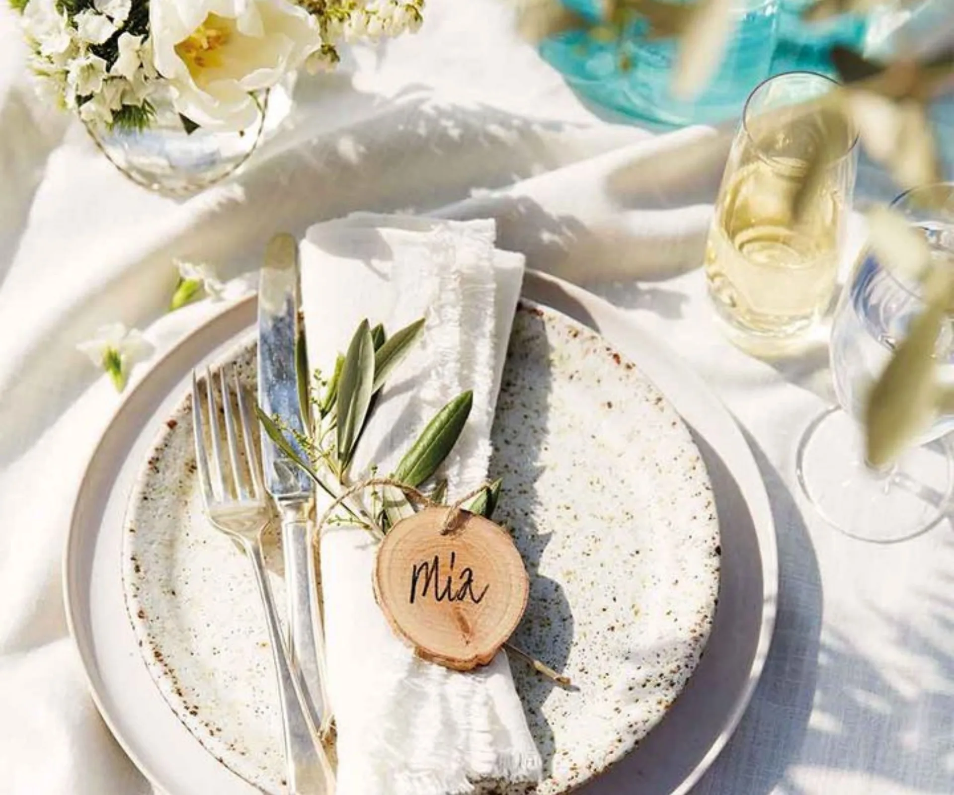 Place setting with speckled plate, utensils, and napkin, decorated with greenery and a wood slice name tag reading "Mia".
