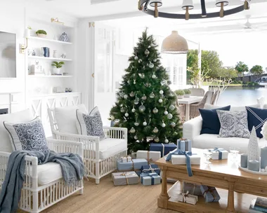 Coastal living room with a Christmas tree, white furniture, blue accents, gifts beneath tree, large windows, and chandelier.