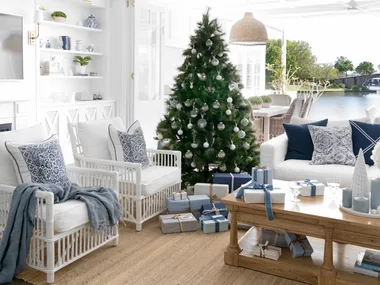 Coastal living room with a Christmas tree, white furniture, blue accents, gifts beneath tree, large windows, and chandelier.