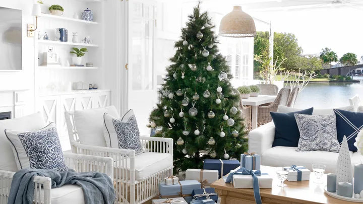 Coastal living room with a Christmas tree, white furniture, blue accents, gifts beneath tree, large windows, and chandelier.