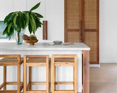 Modern kitchen with wooden stools, white countertop, leafy plant, and woven cabinet doors. Bright and minimalist design.