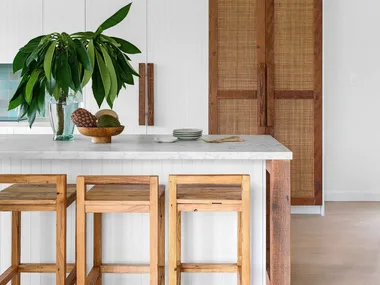 Modern kitchen with wooden stools, white countertop, leafy plant, and woven cabinet doors. Bright and minimalist design.
