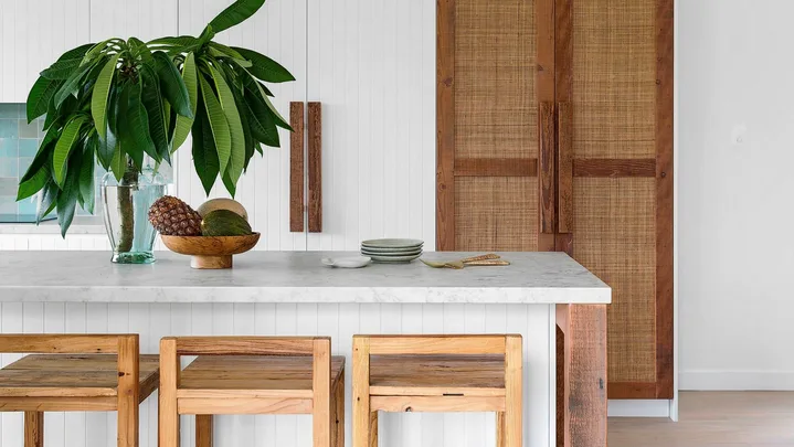 Modern kitchen with wooden stools, white countertop, leafy plant, and woven cabinet doors. Bright and minimalist design.