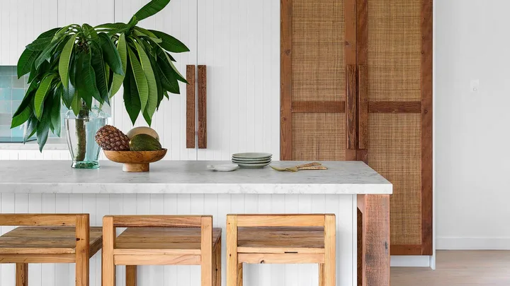 Modern kitchen with wooden stools, white countertop, leafy plant, and woven cabinet doors. Bright and minimalist design.