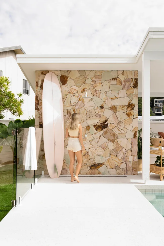 A woman in summer attire stands beside a surfboard by a stone wall near an outdoor pool on a sunny day.
