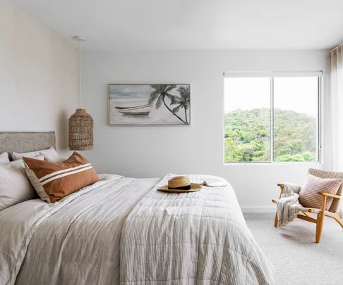 Modern bedroom with a neutral palette, large bed, window view of greenery, and tropical artwork on the wall.