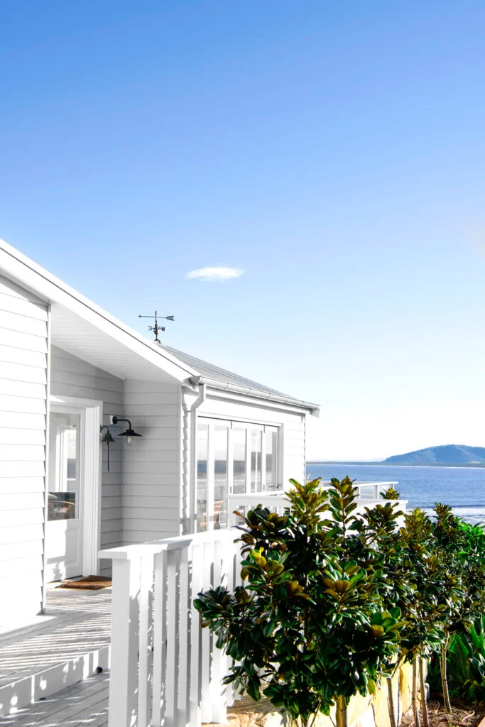 Exterior of a white coastal house with water  views with an evergreen magnolia tree in the foreground.