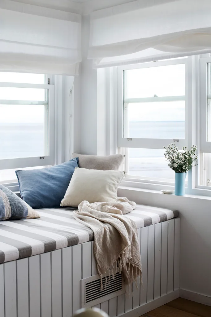 Cozy window seat with striped mattress, blue and beige pillows, a beige throw, and a vase with white flowers by two windows.