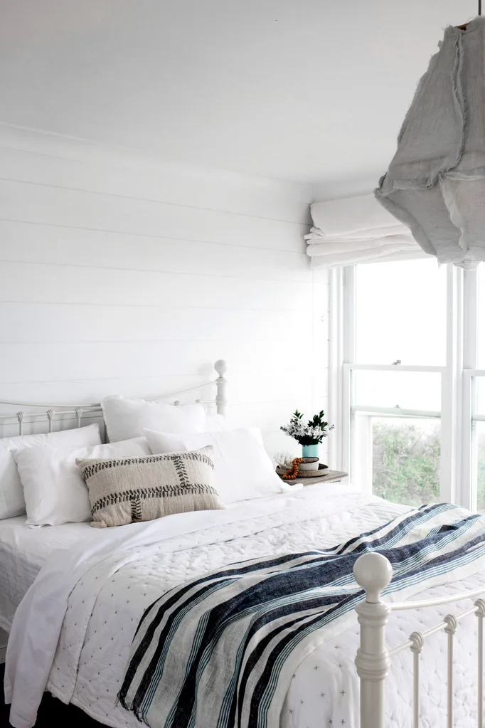 Cozy bedroom with white bedding, striped blanket, and decorative pillows; bright window with light curtains and plant decor.