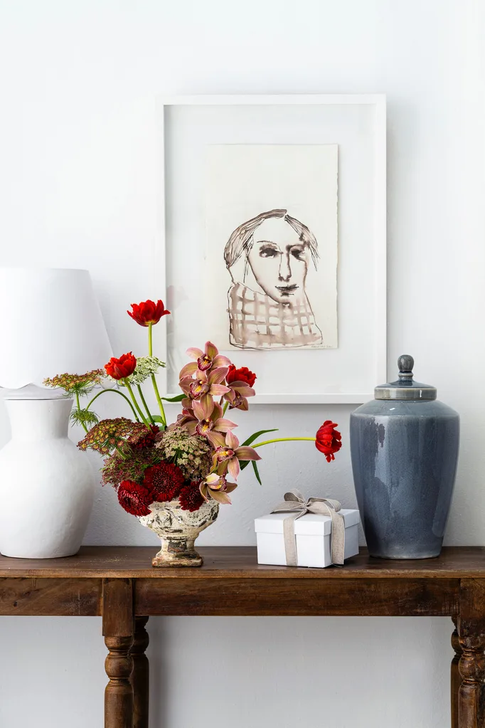 A decorative table with a framed sketch, a white lamp, red flowers, a wrapped gift, and a blue vase.