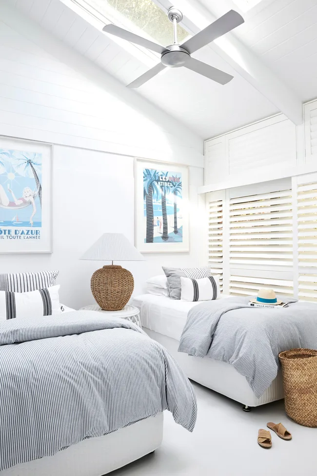 Bright bedroom with two beds, striped bedding, a wicker lamp, framed art, and a ceiling fan.