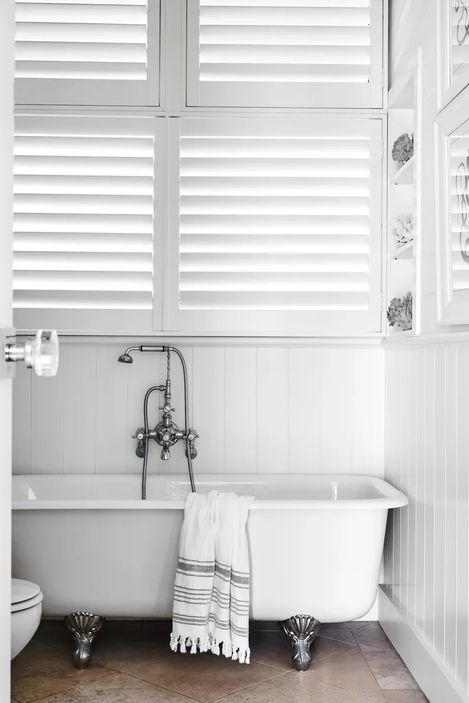 Clawfoot bathtub with silver fixtures, white towel, and shutters in a bright, white bathroom.