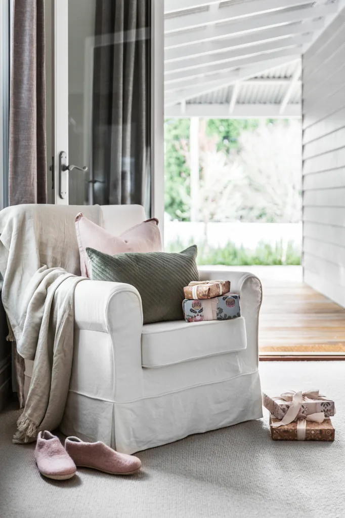 Cozy living room with white armchair, pastel pillows, gift boxes, and slippers by the door.