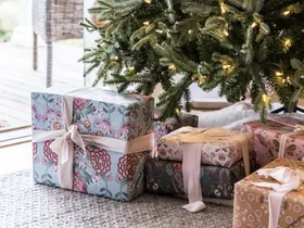 Christmas tree with lights and wrapped gifts in floral paper underneath on a carpeted floor near a window.