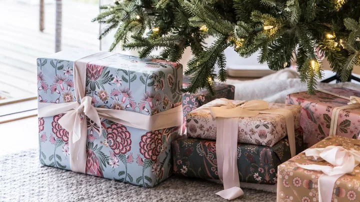 Christmas tree with lights and wrapped gifts in floral paper underneath on a carpeted floor near a window.