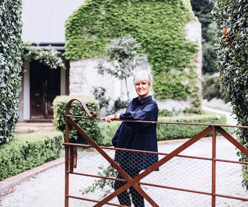 Woman with short gray hair in a dark dress standing by a rustic gate in front of a house covered in greenery.