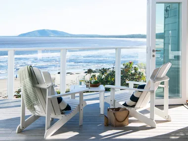 Two white chairs on a beachside deck with a view of the ocean and mountains; glass doors open to a sunny scene.