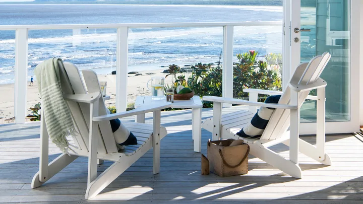 Two white chairs on a beachside deck with a view of the ocean and mountains; glass doors open to a sunny scene.