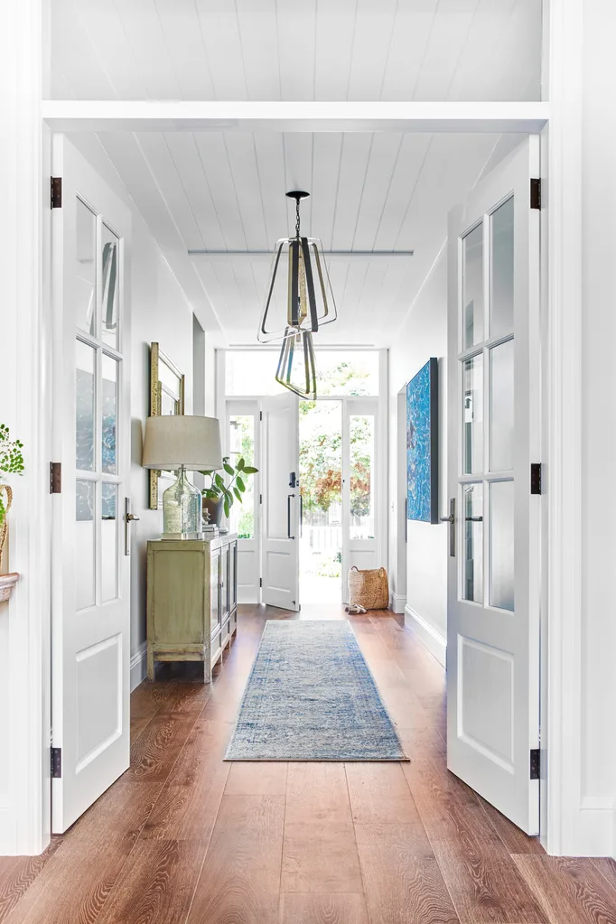 Hallway with wooden floor, blue rug, and modern chandelier; doors open to a bright garden view.