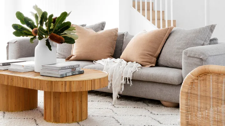 Bright living room with gray sofa, wooden coffee table, and plant decor on a white rug, leading to a stairway with wall art.