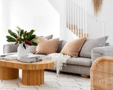 Bright living room with gray sofa, wooden coffee table, and plant decor on a white rug, leading to a stairway with wall art.